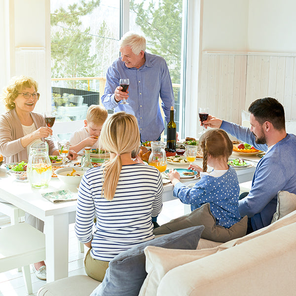 Famiglia che si raccoglie attorno a un tavolo per un pasto conviviale, brindando con bicchieri di vino o succo in una stanza luminosa.